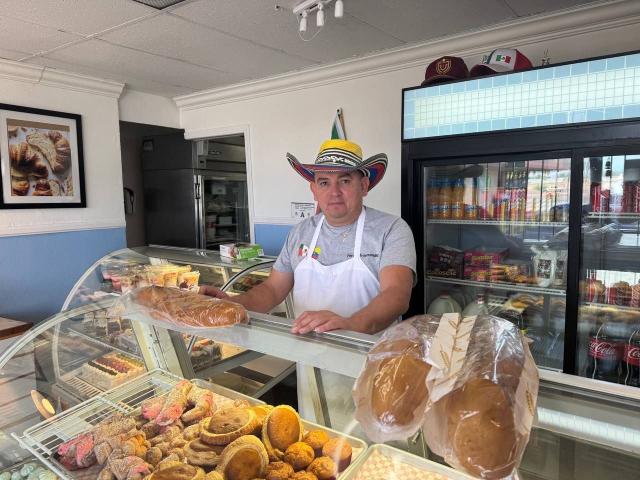 Man wearing traditional hat and apron behind bakery counter with assorted bread and pastries.